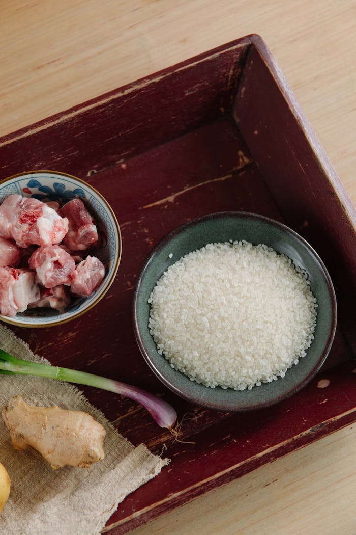 Overhead view of raw meat, ginger, and rice on a wooden tray, ready for cooking.